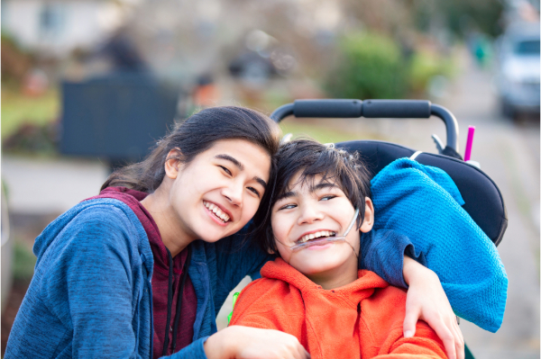 A boy with dark hair who uses a wheelchair is embraced by a young woman. They are both smiling. A boy with dark hair who uses a wheelchair is embraced by a young woman. They are both smiling.