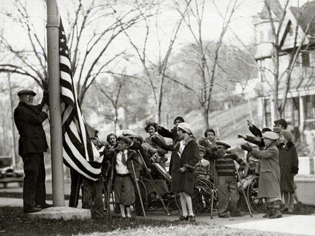 A man raises a large flag as many children try to help and salute at the same time. Many of the children are using wheelchairs, crutches, and braces for support as they stand on the lawn of a new building.